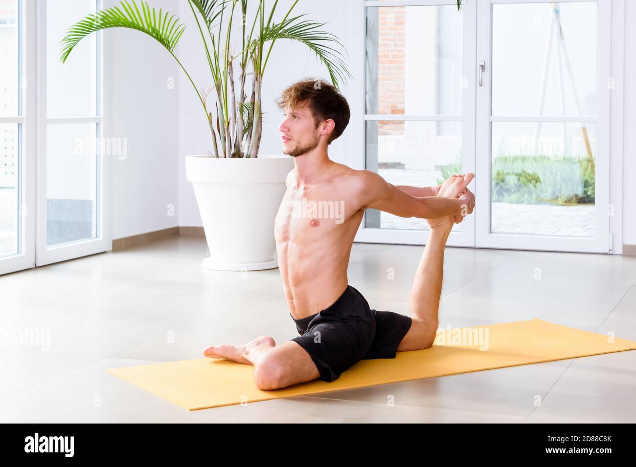Fit jungen Mann tun eine Eka Pada Kapotasna Yoga-Pose, um seine Hüften, Leistengegend und Kernmuskeln in einem High-Key-Fitnessstudio mit Copyspace in einer Gesundheit und fitne strecken Stockfoto