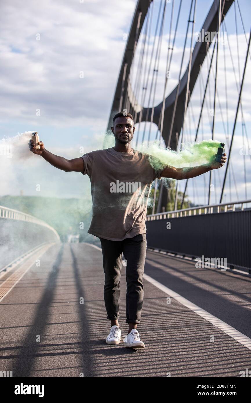 Junger schwarzer Mann, der mit Rauchbomben in ausgestreckten Armen geht. Volle Länge. Vertikal. Stockfoto