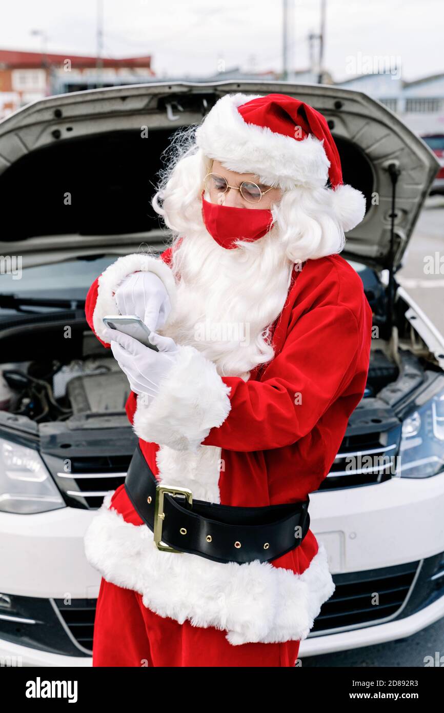 Porträt des Weihnachtsmannes, der etwas in sein Handy schreibt Besorgt und mit der Haube seines Autos geöffnet, weil Sein Auto ist kaputt gegangen und er kann nicht d Stockfoto