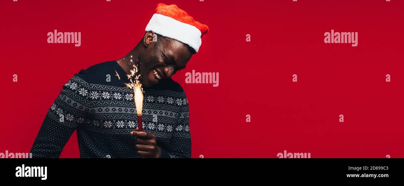 Fröhlicher junger afrikanischer Mann mit Wunderkerzen auf rotem Hintergrund. Jungen Kerl trägt einen santa hut mit Feuerwerk. Stockfoto