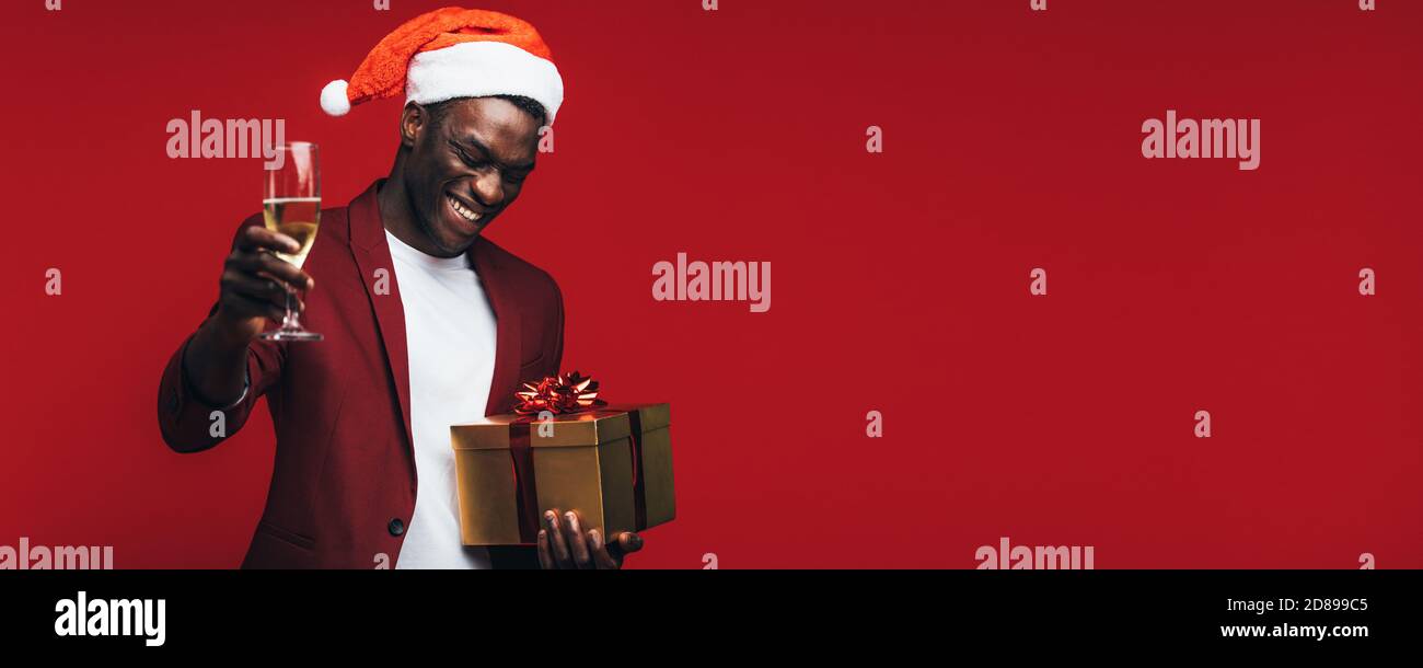 Afrikanischen Mann, santa hut mit Champagner und Geschenk Box gegen roten Hintergrund. Glückliche junge schwarze Kerl genießen Heiligabend. Stockfoto