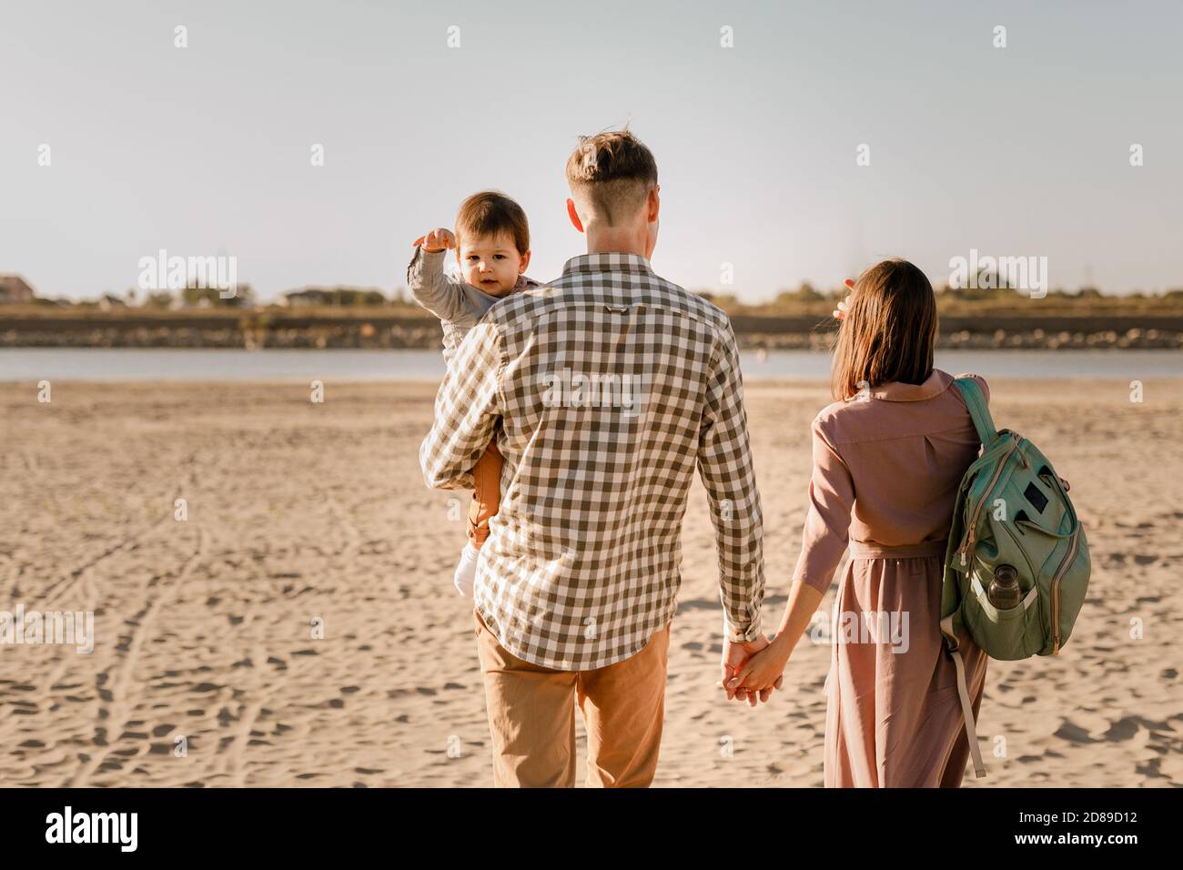 Glückliche Familie zu Fuß am Sandstrand des Flusses. Vater, Mutter hält Baby Sohn an den Händen und geht zusammen. Rückansicht. Familienbande-Konzept Stockfoto