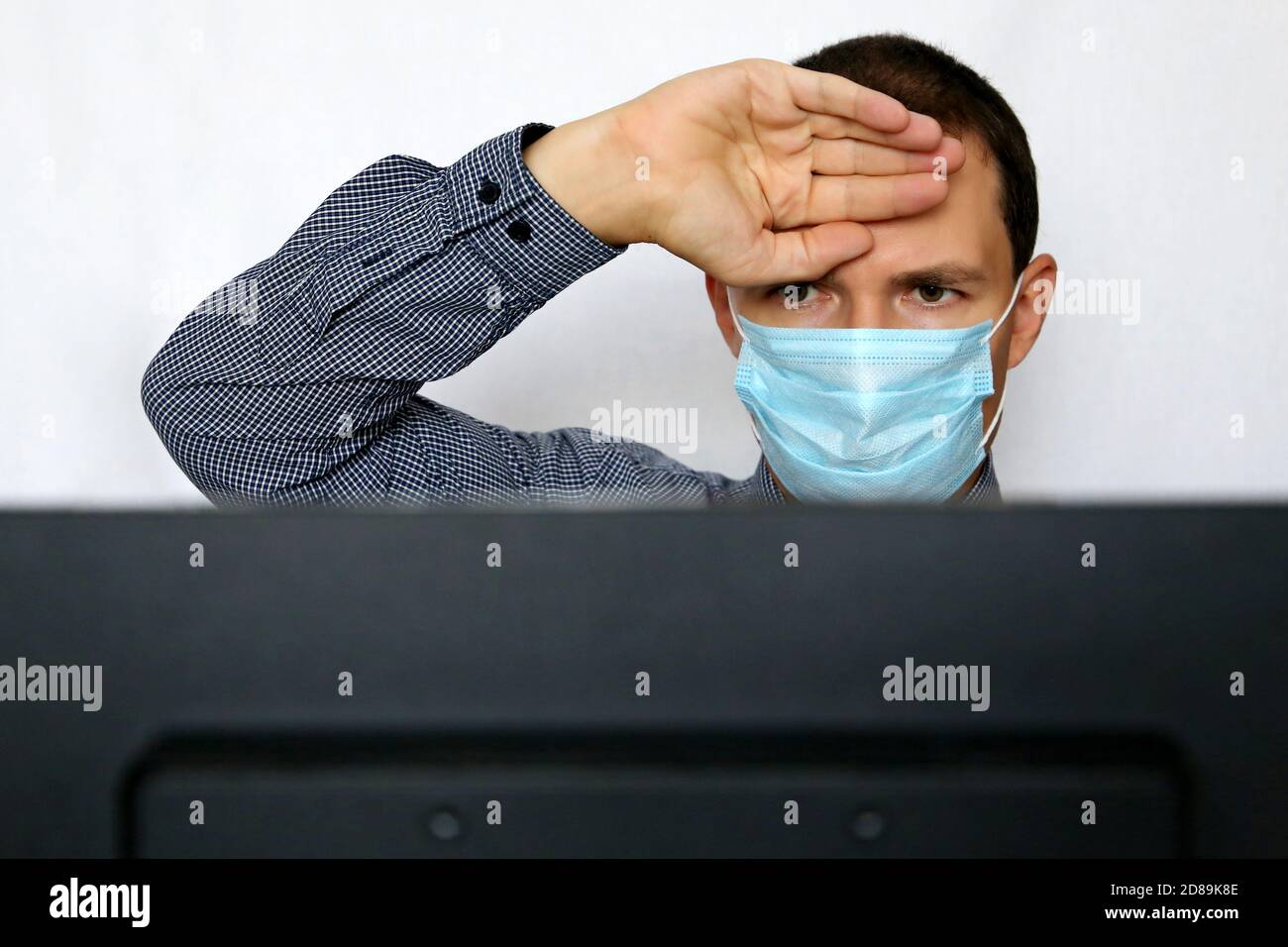 Kranker Mann in Gesichtsmaske, der seine Stirn am PC-Monitor hält. Sicherheit am Arbeitsplatz während einer Coronavirus-Pandemie Stockfoto