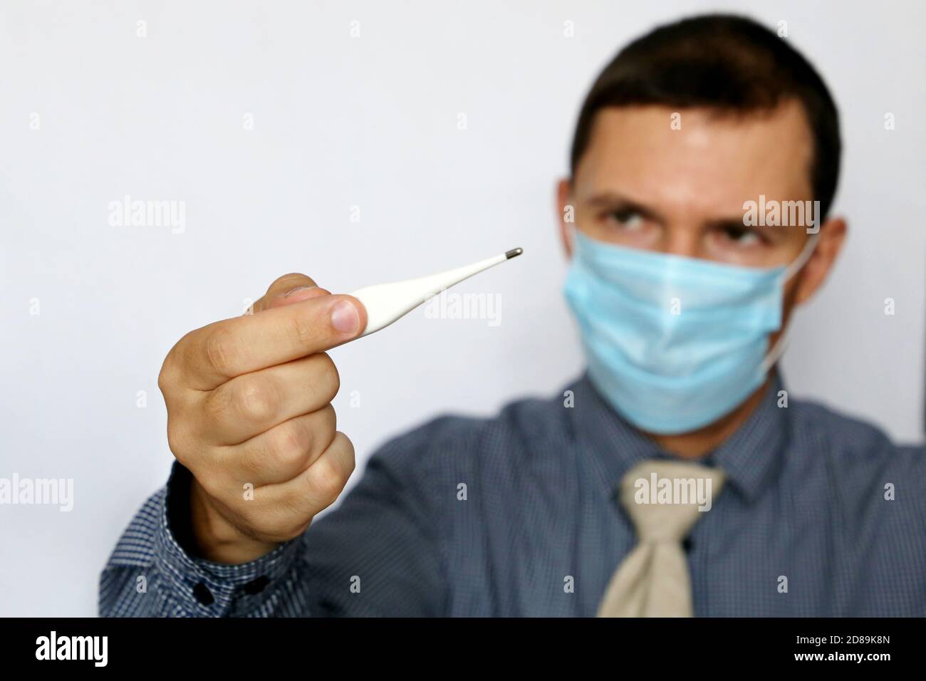Mann in Gesichtsmaske und Bürokleidung am PC-Monitor schaut auf Digitalthermometer in der Hand. Hohe Körpertemperatur, Sicherheit bei der Arbeit während des Coronavirus Stockfoto