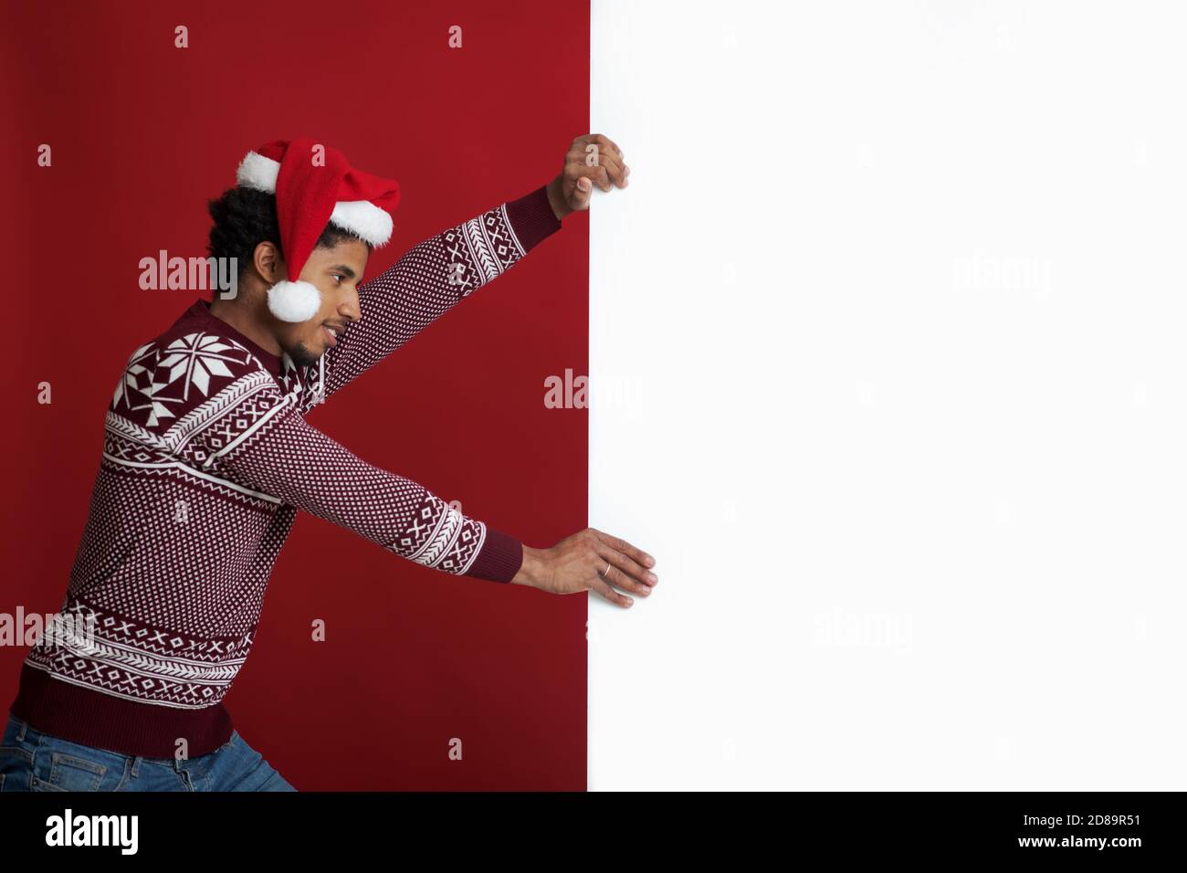 afroamerikanischer junger Mann in Pullover und weihnachtsmütze schiebt Mit Aufwand weißes Banner mit Leerzeichen Stockfoto