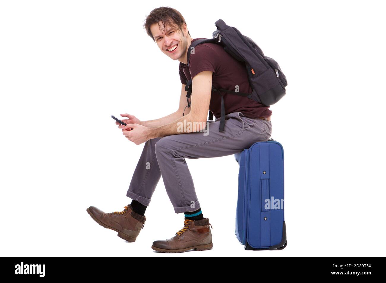 In voller Länge Seite Porträt von glücklichen jungen Reisenden mit Handy Und auf dem Koffer sitzen Stockfoto