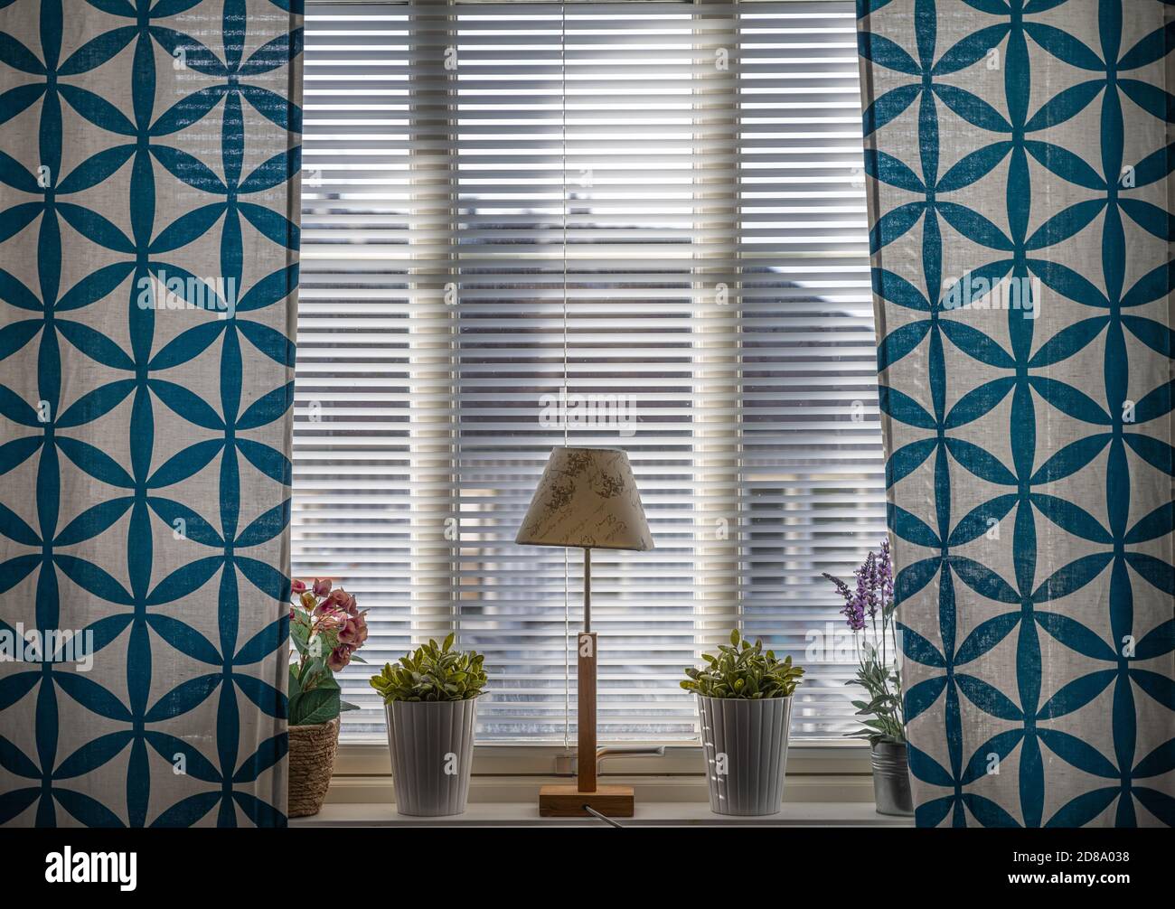 Ein Fenster mit blauen Vorhängen, einer Lampe und Blumen. Schönes Licht kommt durch die Jalousien Stockfoto