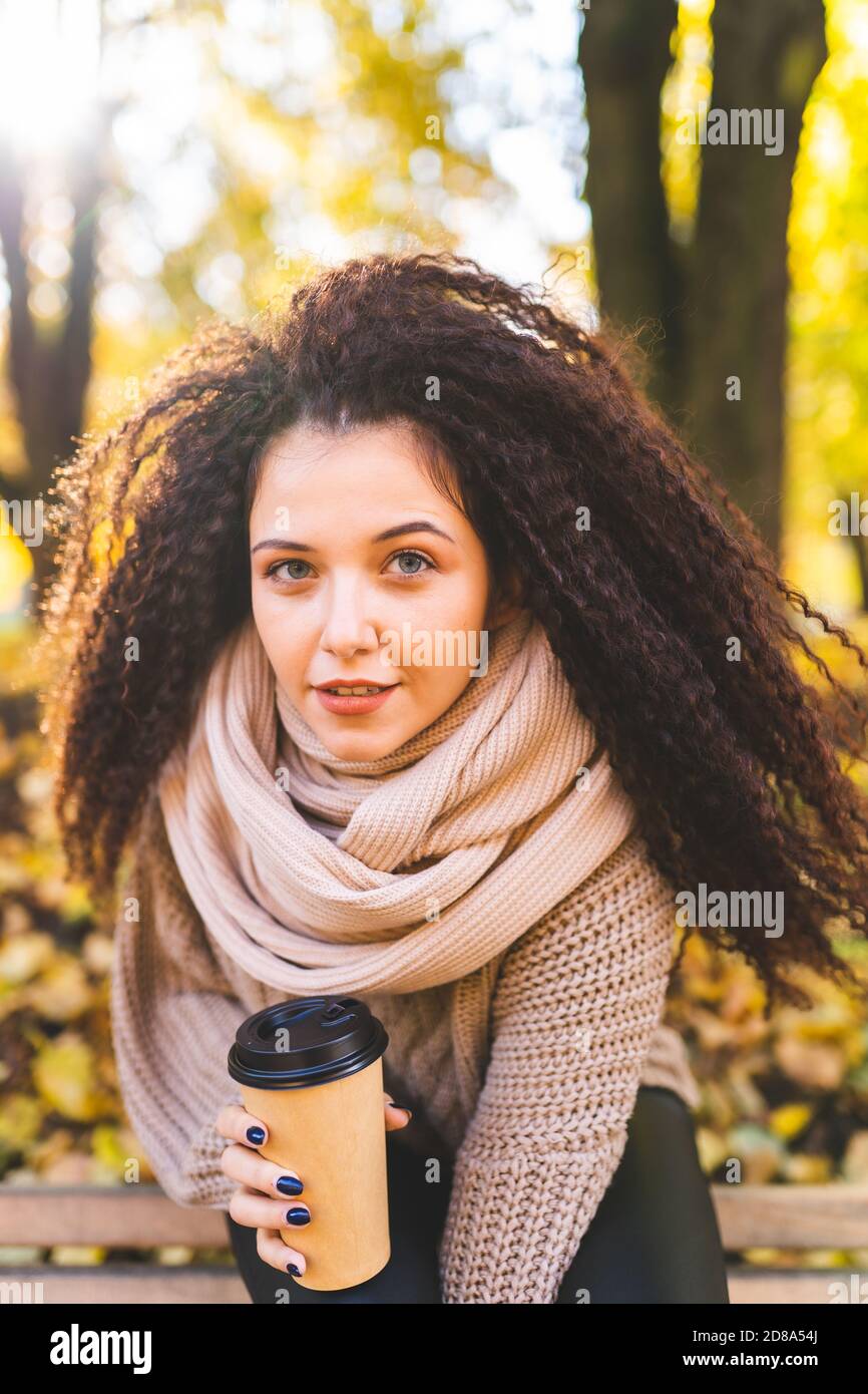 Herbstkonzept - lockige Frau trägt warmen Pullover und Schal trinken Kaffee in Parkbank unter Herbstlaub in sonnigen Tag. Stockfoto