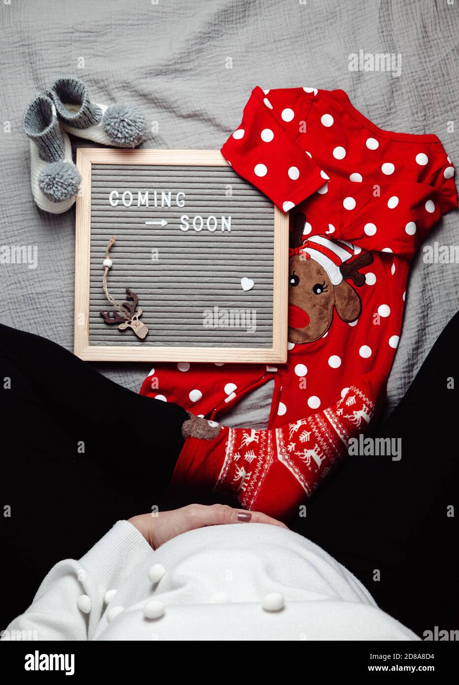 Schwangere Frau sitzt mit einem Coming Soon Baby Ankündigungszeichen. Demnächst Weihnachtskonzept. Schwangerschaftsbauch. Leerzeichen. Stockfoto