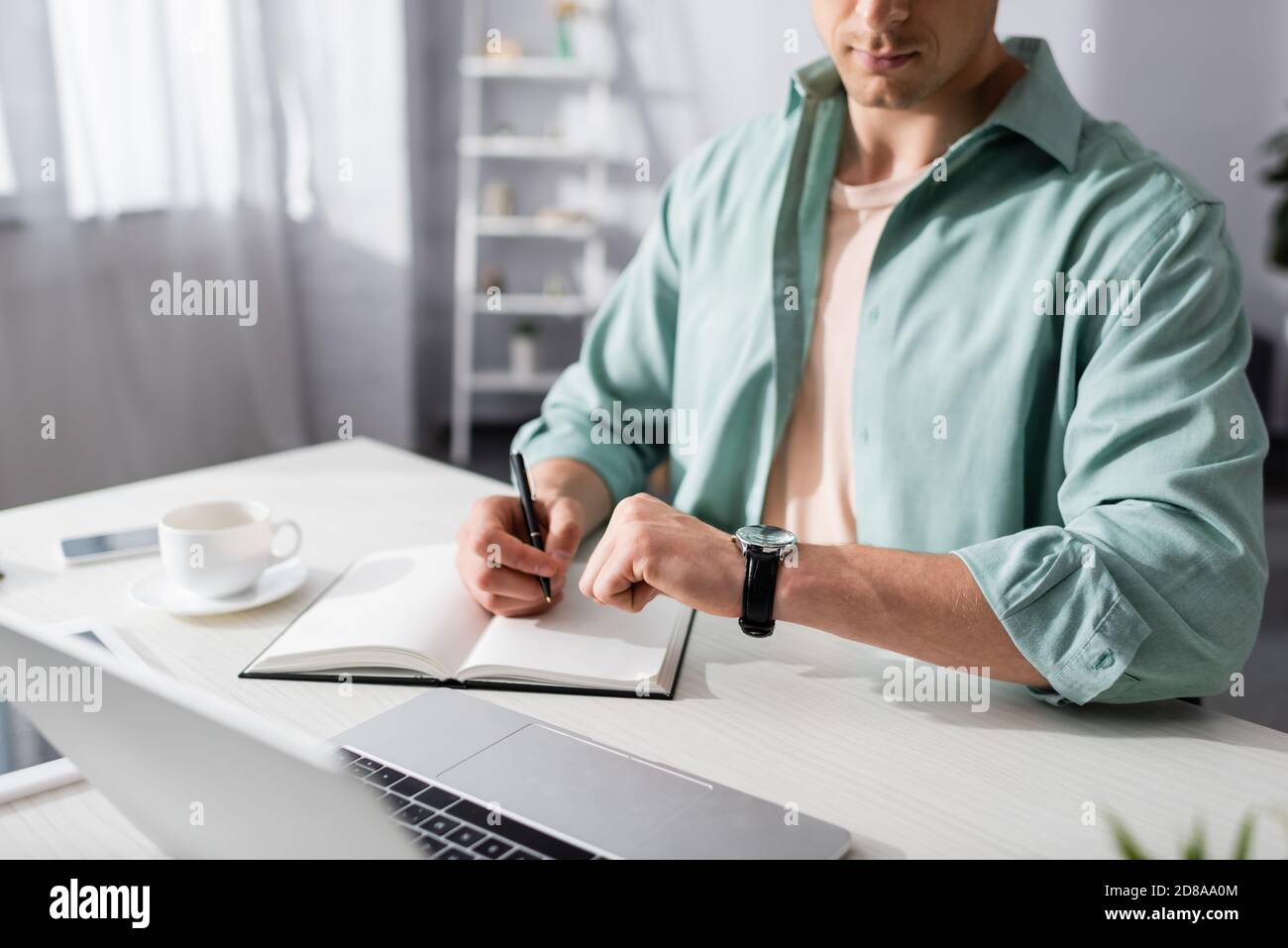 Beschnittene Ansicht des Freiberuflers, der Zeit auf der Armbanduhr während der Arbeit mit Notebook und Laptop zu Hause überprüft, Konzept des Zeitmanagements Stockfoto