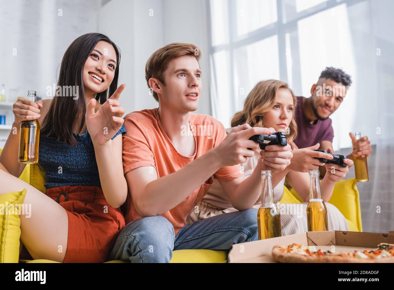 KIEW, UKRAINE - 28. JULI 2020: Aufgeregt asiatische Frau hält Bier und zeigt mit dem Finger in die Nähe von Freunden spielen Videospiel Stockfoto