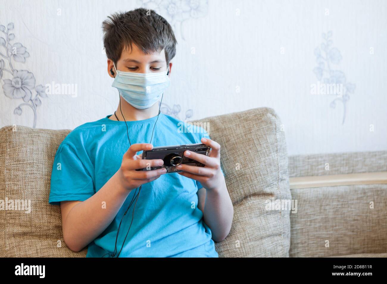Teenager trägt Gesichtsmaske und hört die Musik mit Ohrhörern Und Smartphone, während Sie zu Hause sitzen Stockfoto
