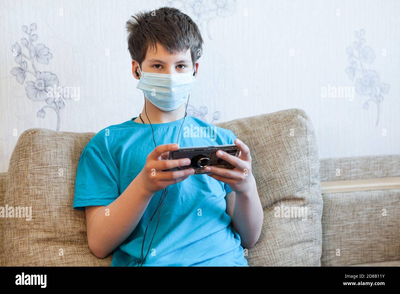 Pre-teen Junge trägt medizinische Maske Musik hören mit Kopfhörern und Handy während man im heimischen Zimmer auf der Couch sitzt Stockfoto