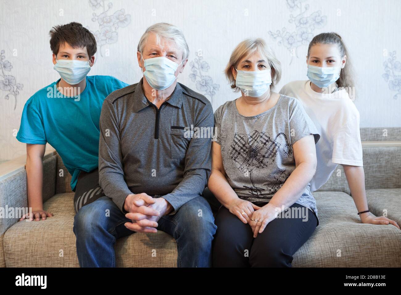 Familie, die während einer Coronavirus-Pandemie zu Hause bleibt, Sicherheitsmaske trägt, zusammen auf einer Couch sitzt und die Kamera anschaut Stockfoto