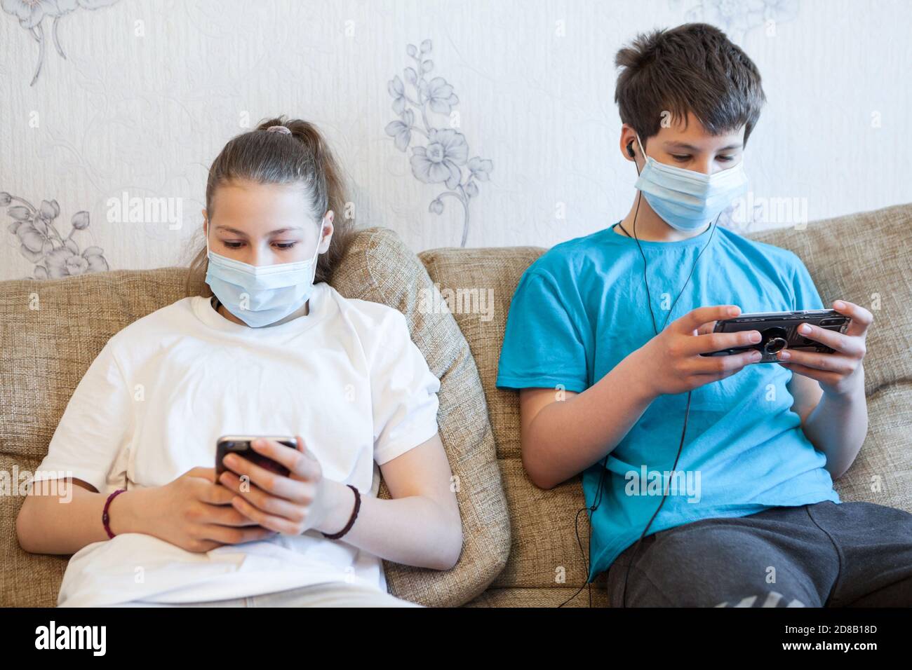 Jugendliche Jungen und Mädchen mit Smartphones für Online-Gespräche, Kinder tragen One-Use-Masken, sitzen auf dem Sofa Stockfoto