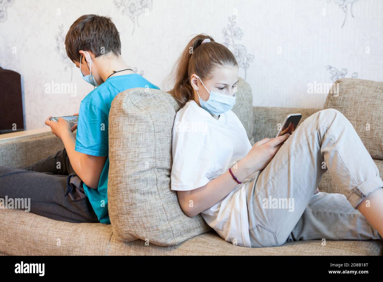 Kinder spielen mit Handys auf dem Sofa, tragen medizinische Masken beide Stockfoto