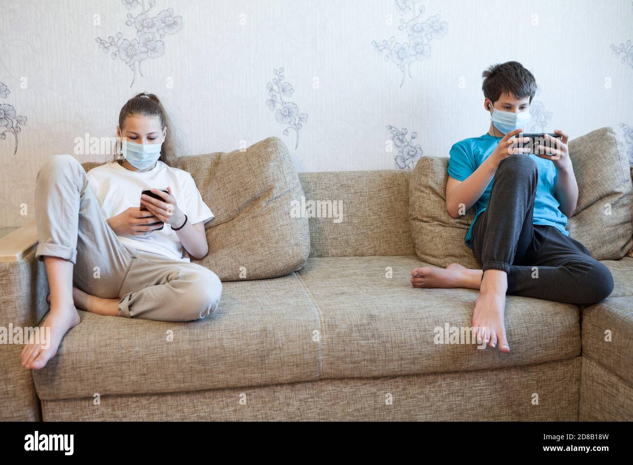 Zwei Kinder halten Abstand, während sie Smartphones benutzen, Kinder tragen Gesichtsmaske zu Hause während der Coronavirus-Pandemie Stockfoto