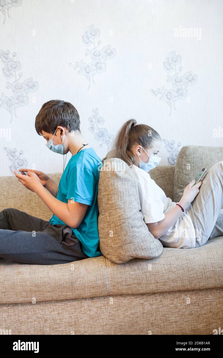 Zwei Kinder vor dem Teenager spielen online mit Mobiltelefonen, während sie zu Hause sitzen, Kinder tragen Gesichtsmasken und sitzen zurück auf der Couch Stockfoto