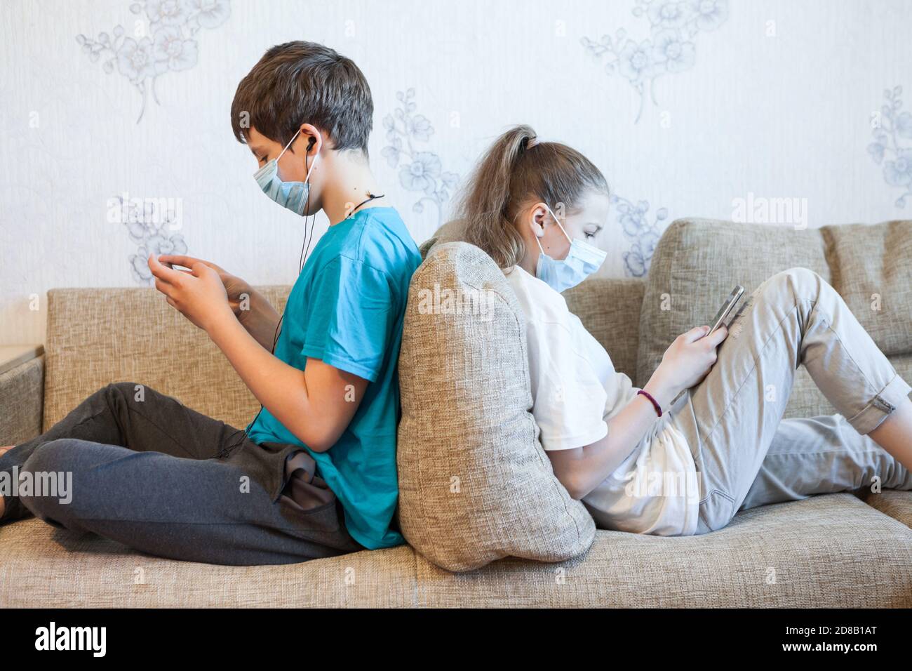 Schwester und Bruder mit ihren Smartphones, während sie zu Hause auf der Couch sitzen und Gesichtsmasken tragen Stockfoto