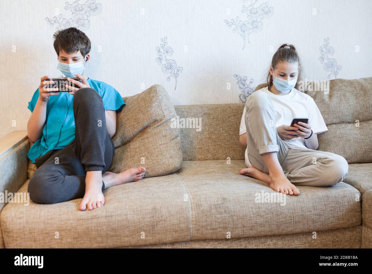 Jugendliche Jungen und Mädchen spielen online mit Smartphones, während sie zu Hause bleiben, Kinder tragen medizinische Masken, Abstand halten Stockfoto