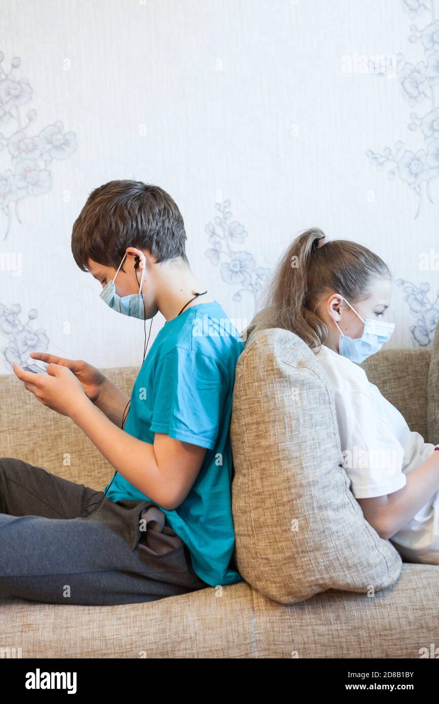 Kleine Kinder spielen mit ihren Handys auf einer Couch und tragen medizinische Masken Stockfoto