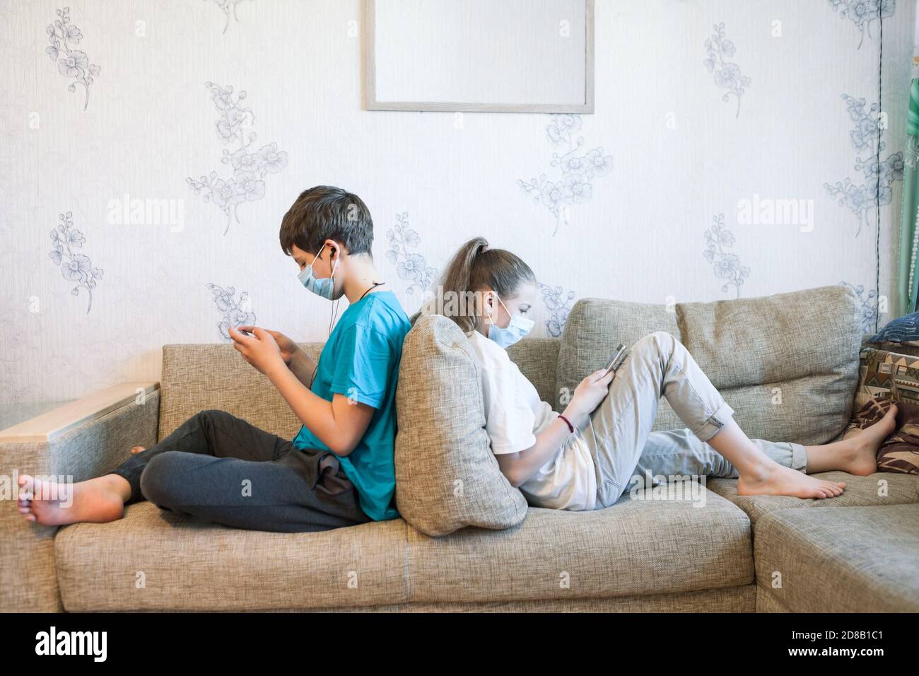 Pre-teen Schwester und Bruder online mit ihren Handys spielen, während zu Hause sitzen, Kinder tragen Sicherheitsmasken, sitzen zurück auf dem Sofa Stockfoto