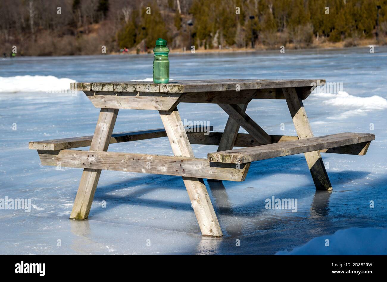Ein hölzerner Picknicktisch auf einem gefrorenen See. Eine grüne Wasserflasche sitzt auf dem Tisch. Sonniger Tag. Hinweis Horizont ist nicht schief, dies ist die Form von Th Stockfoto