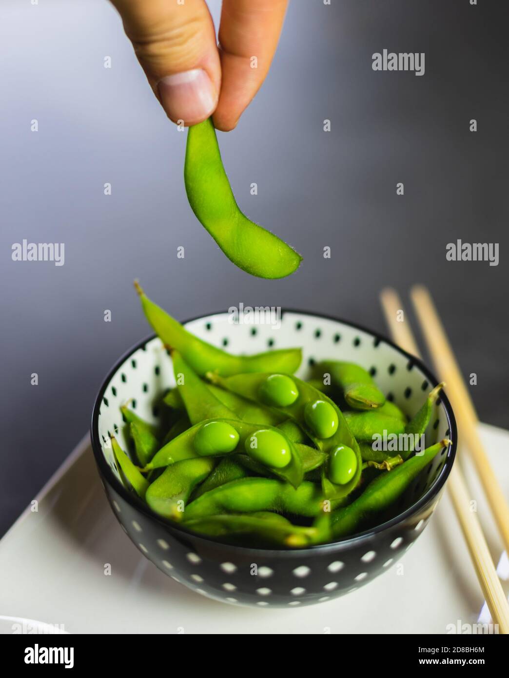 Hand nimmt eine edamame Bohne aus einer Schüssel. Frische grüne Sojabohnen in einer Schüssel mit Essstäbchen. Stockfoto