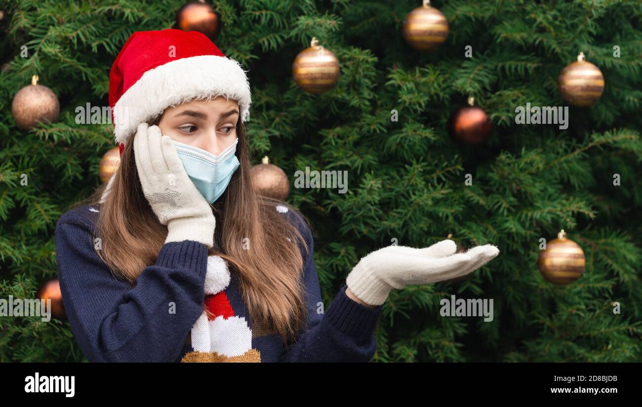 Weihnachten ohne Geschenk. Nahaufnahme Porträt einer Frau, die einen weihnachtsmann Hut und medizinische Maske mit Emotionen trägt. Vor dem Hintergrund eines Weihnachtsstr Stockfoto