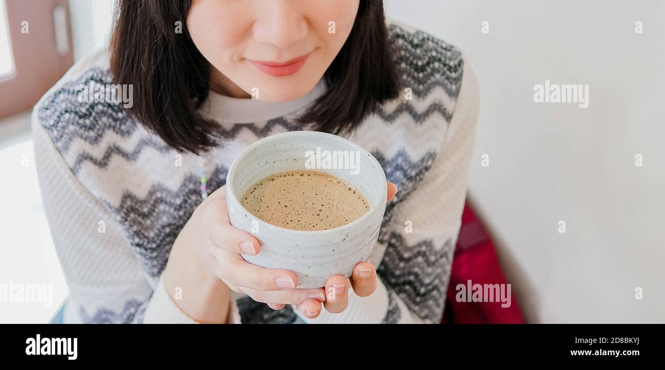 Junge Erwachsene asiatische Frau trägt einen Strickpullover genießen Sie ein heißes Getränk am Morgen in einem weißen Raum Hintergrund. Ein glückliches lächelndes Mädchen, das sich drinnen entspannt. Mit Kopierbereich. Stockfoto