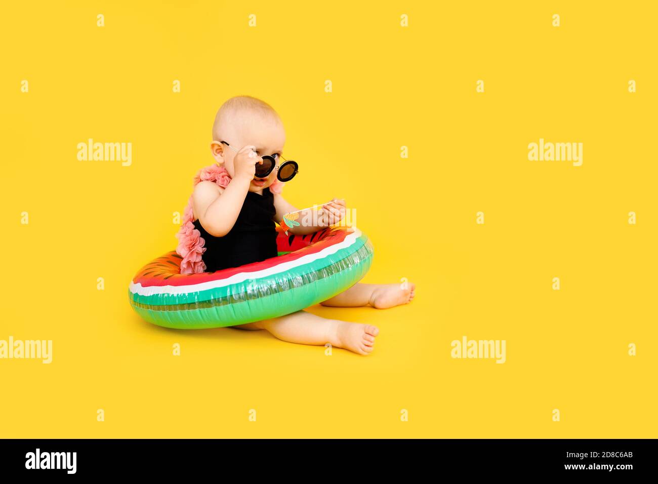 Baby Mädchen in schwarz und rosa Badeanzug hält Wassermellon aufblasbaren Pool schweben auf gelbem Hintergrund mit Kopie Raum. Stockfoto