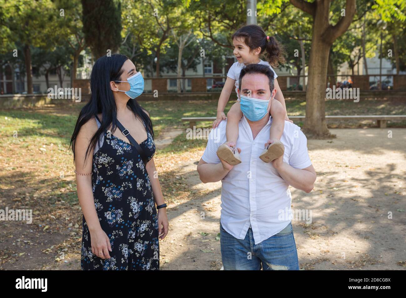 Ein kaukasischer Vater hält sein 2-jähriges Mestizo-Mädchen in einem Park in barcelona CI, während die lateinische Mutter sie anschaut. Die Eltern tragen ein schützendes Mas Stockfoto