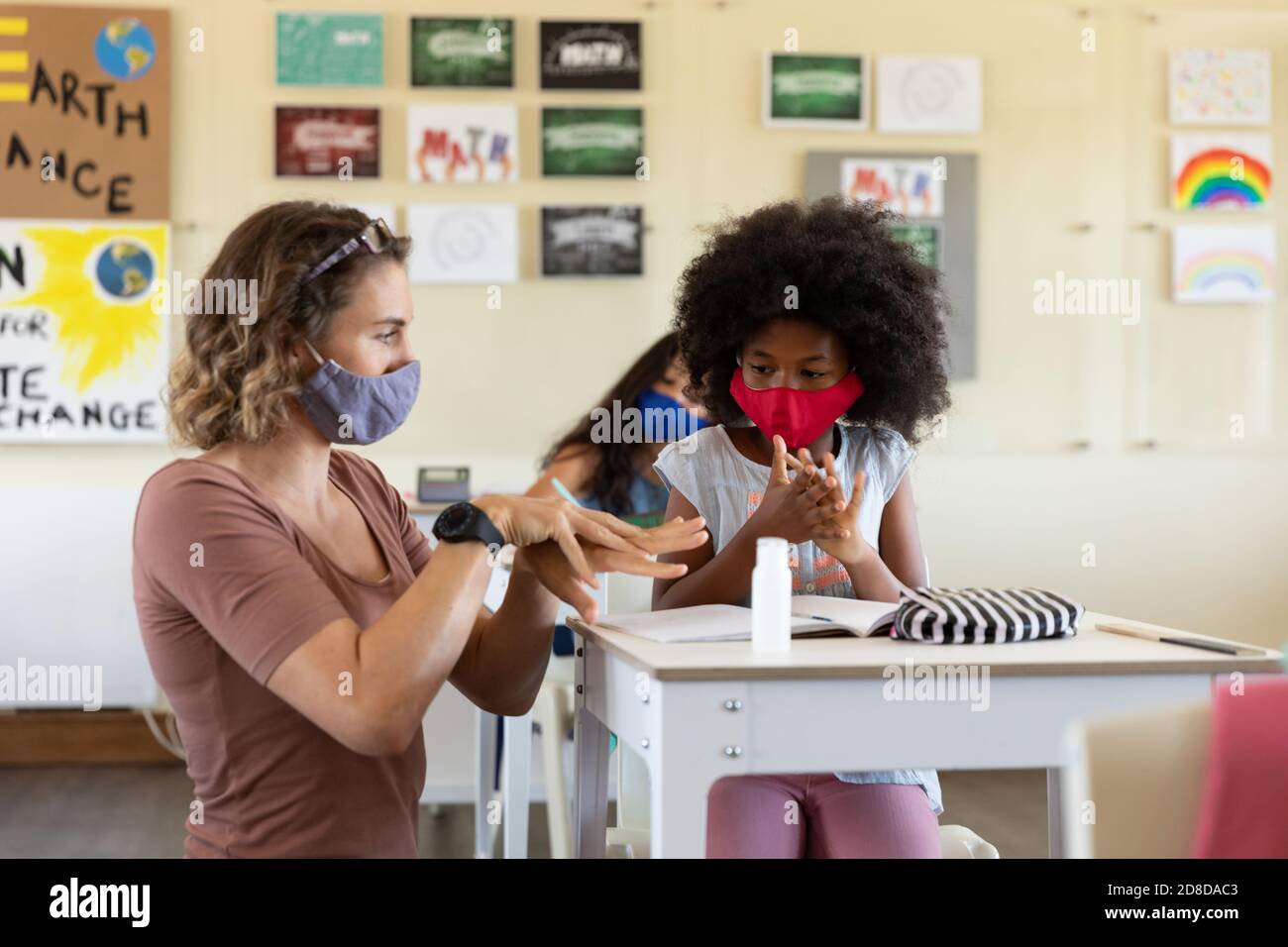 Weibliche Lehrerin trägt Gesichtsmaske hilft einem Mädchen zu desinfizieren Ihre Hände in der Klasse Stockfoto