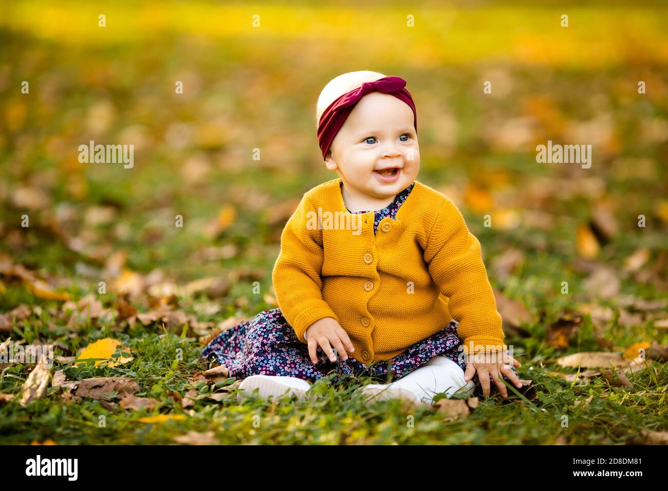 Baby-Mädchen in yelloy Jacke und roten Stirnband sitzen auf dem Gras, spielen in den Herbstblättern. Stockfoto