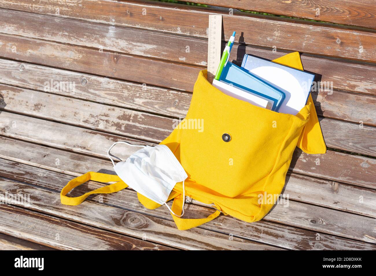 Gelber Textilrucksack mit Schulbedarf und Büchern auf einer Bank im Freien. Medizinische Maske, Virenschutz. Neues Schuljahr, zurück zur Schule, Pause. Ne Stockfoto