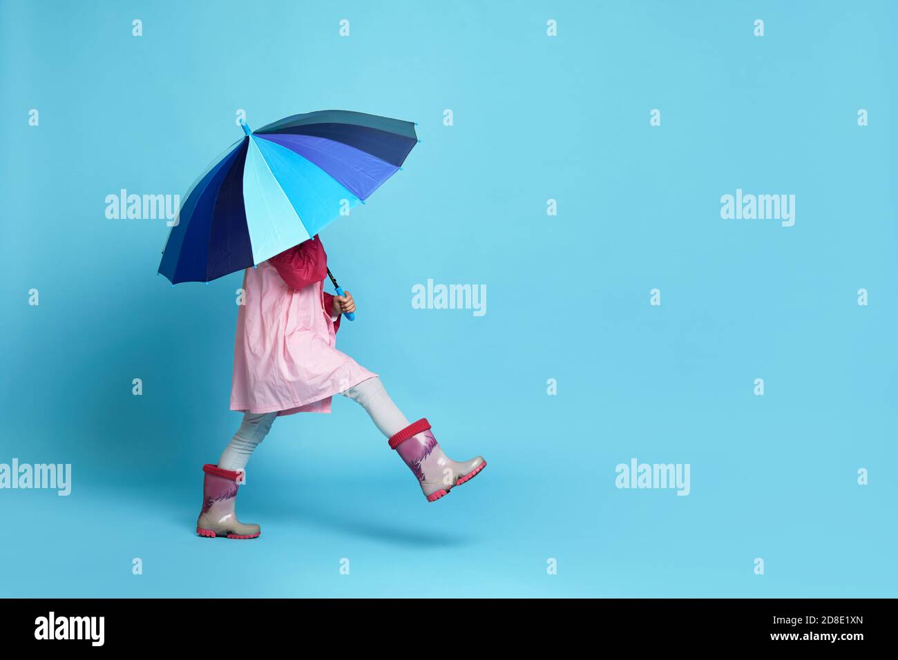 Happy Little Kind Mädchen mit blauem Regenschirm in rosa regen Mantel und Gummistiefel auf blauem Hintergrund. Copy Raum für Text Stockfoto