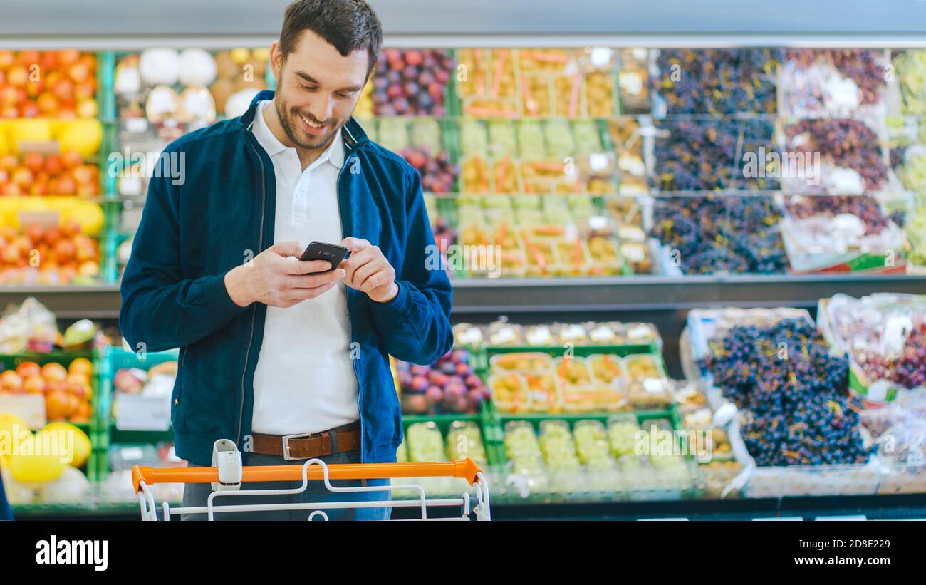 Im Supermarkt: Handsome man benutzt Smartphone, während er in der Frischwarenabteilung des Stores steht. Mann in Internet-Surfen auf seinem eingetaucht Stockfoto