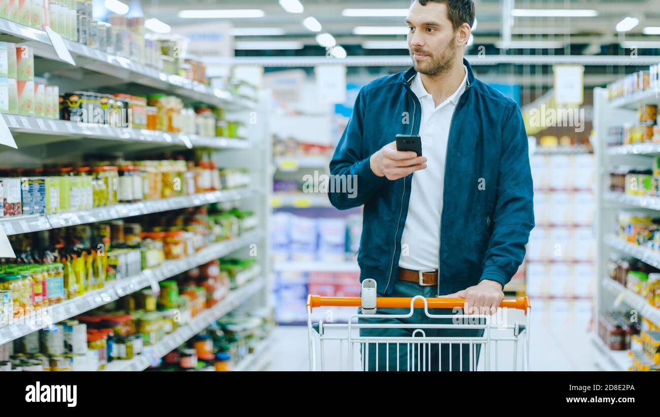 Im Supermarkt: Handsome man nutzt Smartphone und schaut auf den Nährwert der Konserven. Er steht mit dem Warenkorb in Konserven Stockfoto