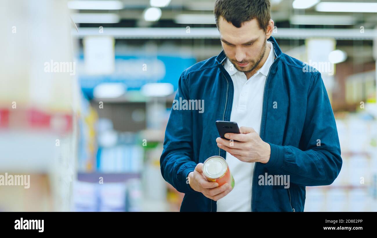 Im Supermarkt: Handsome man nutzt Smartphone, um den Nährwert der Konserven zu überprüfen und zu kaufen. Er steht mit dem Einkaufswagen in Canned Stockfoto