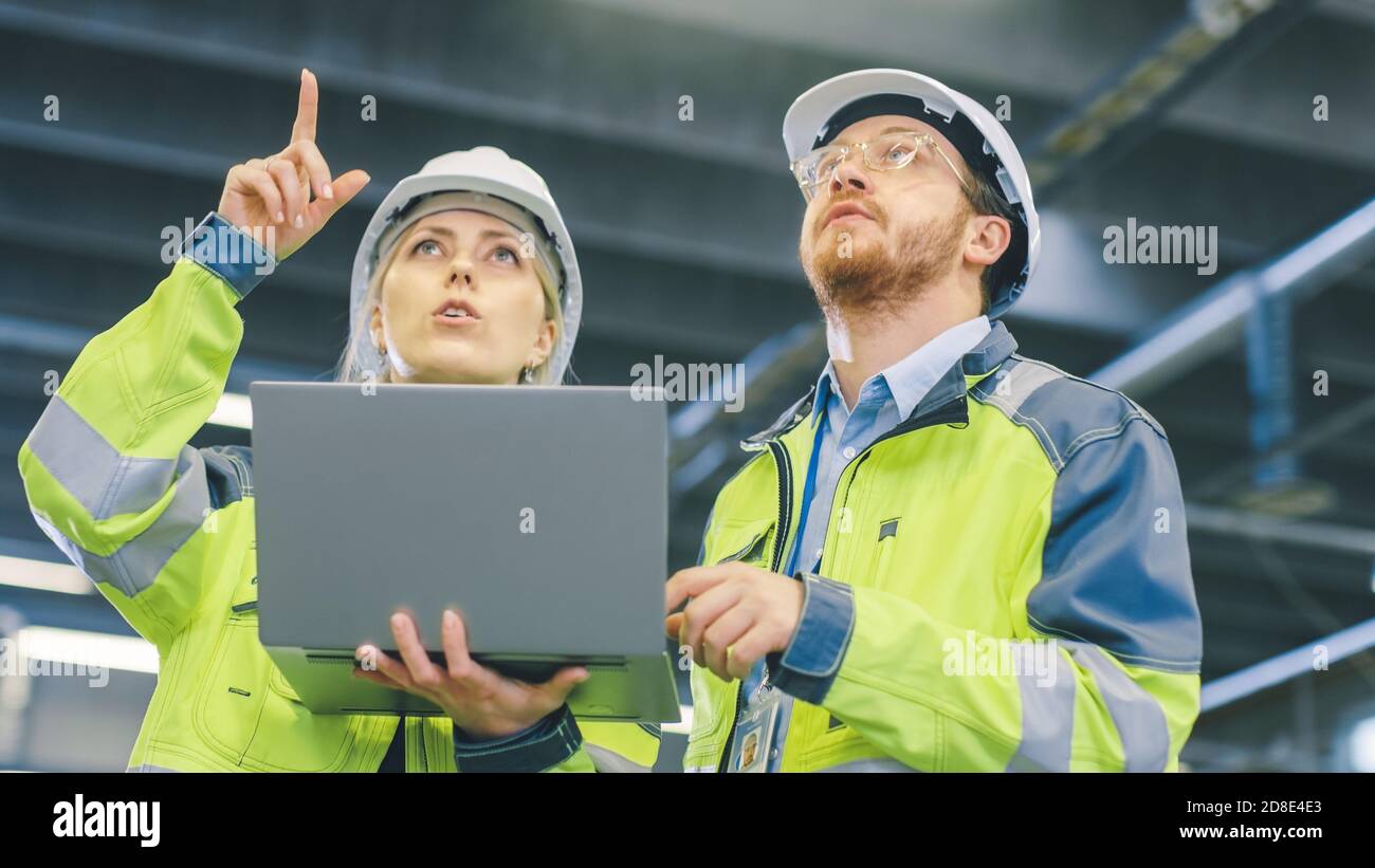 Männliche und weibliche Industrieingenieure arbeiten in einer Fertigungsanlage, sie diskutieren Projekt, zeigen in die Richtung der Maschinen, während Laptop verwenden Stockfoto