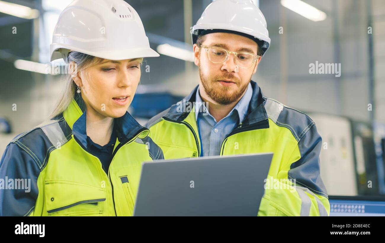 Männliche und weibliche Industrieingenieure arbeiten in einer Fertigungsanlage, sie diskutieren Projekt, zeigen in die Richtung der Maschinen, während Laptop verwenden. Stockfoto