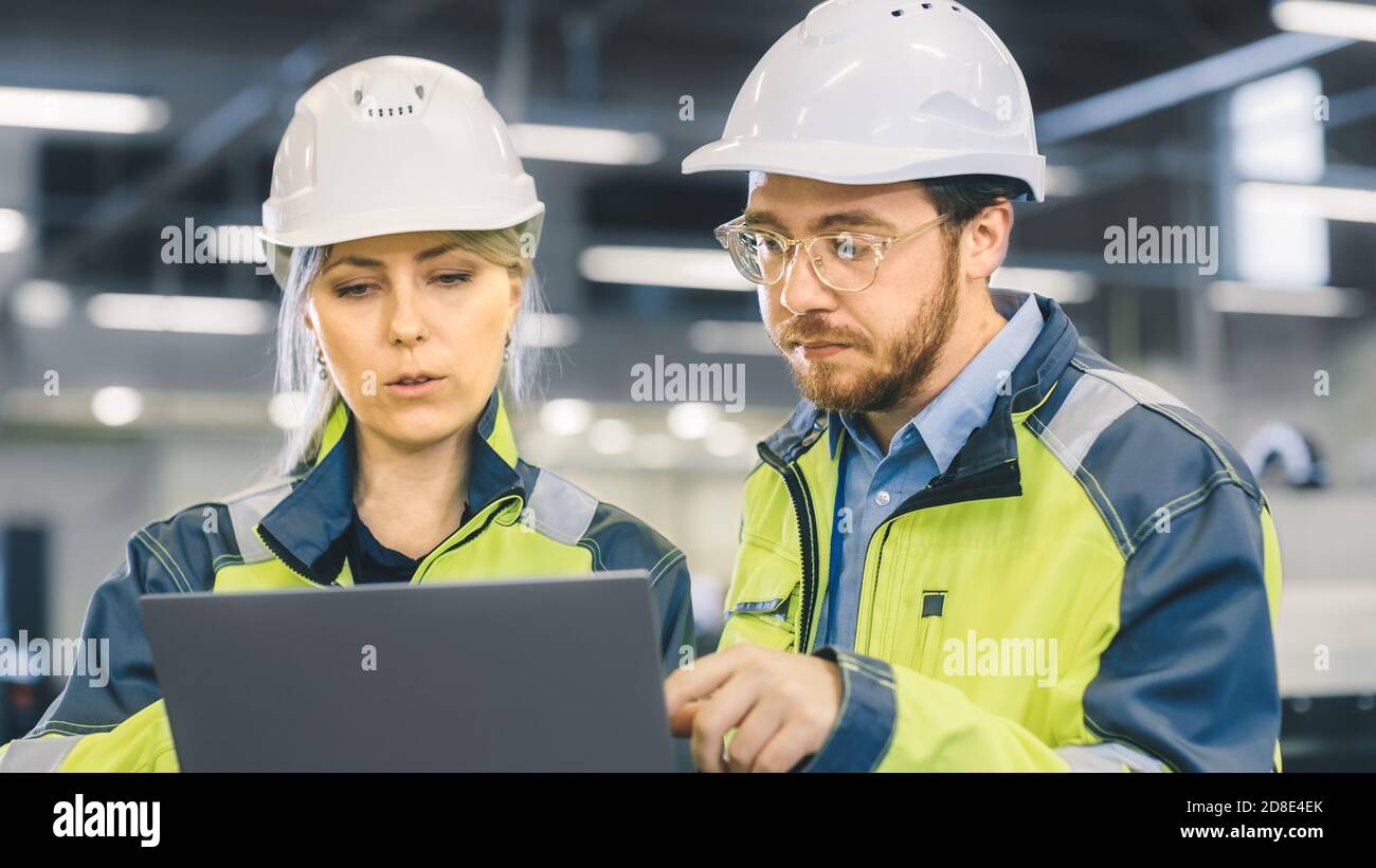 Männliche und weibliche Industrieingenieure arbeiten in einer Fertigungsanlage, sie diskutieren Projekt, zeigen in die Richtung der Maschinen, während Laptop verwenden. Stockfoto