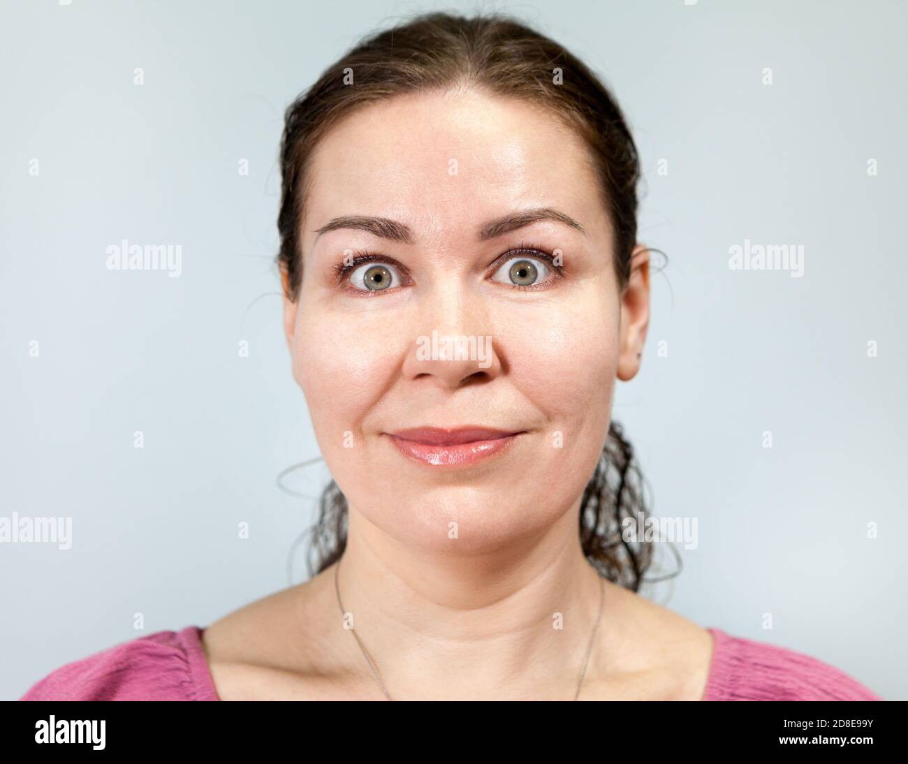 Erwachsene Frau mit einem Lächeln und weit geöffneten Augen, Porträt auf grauem Hintergrund, Emotionen-Serie Stockfoto
