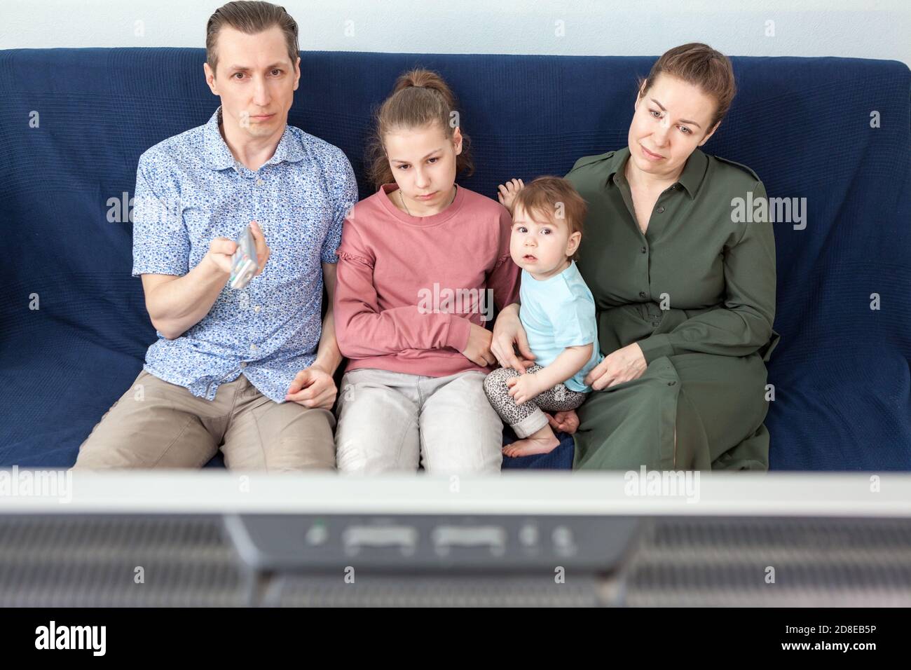 Familie mit Vater, Mutter, Teenager-Tochter und Kleinkind, die TV-Set beobachten, während sie auf dem Sofa im heimischen Zimmer sitzen und zu Hause bleiben Stockfoto