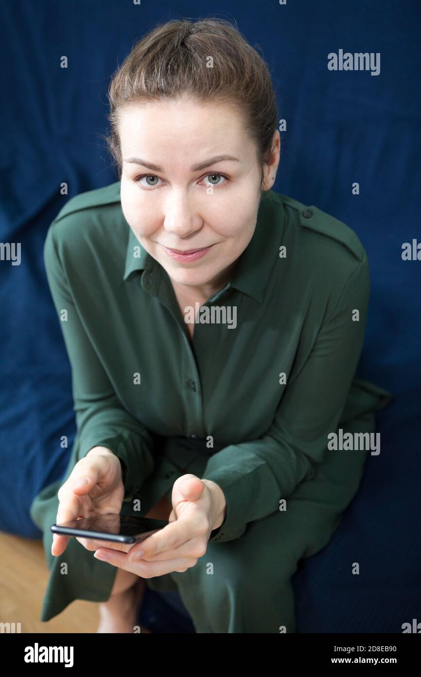 Porträt eines erwachsenen kaukasischen lächelnde Frau in grünem Kleid sitzt auf einer Couch mit Smartphone zu Hause, Blick auf die Kamera Stockfoto
