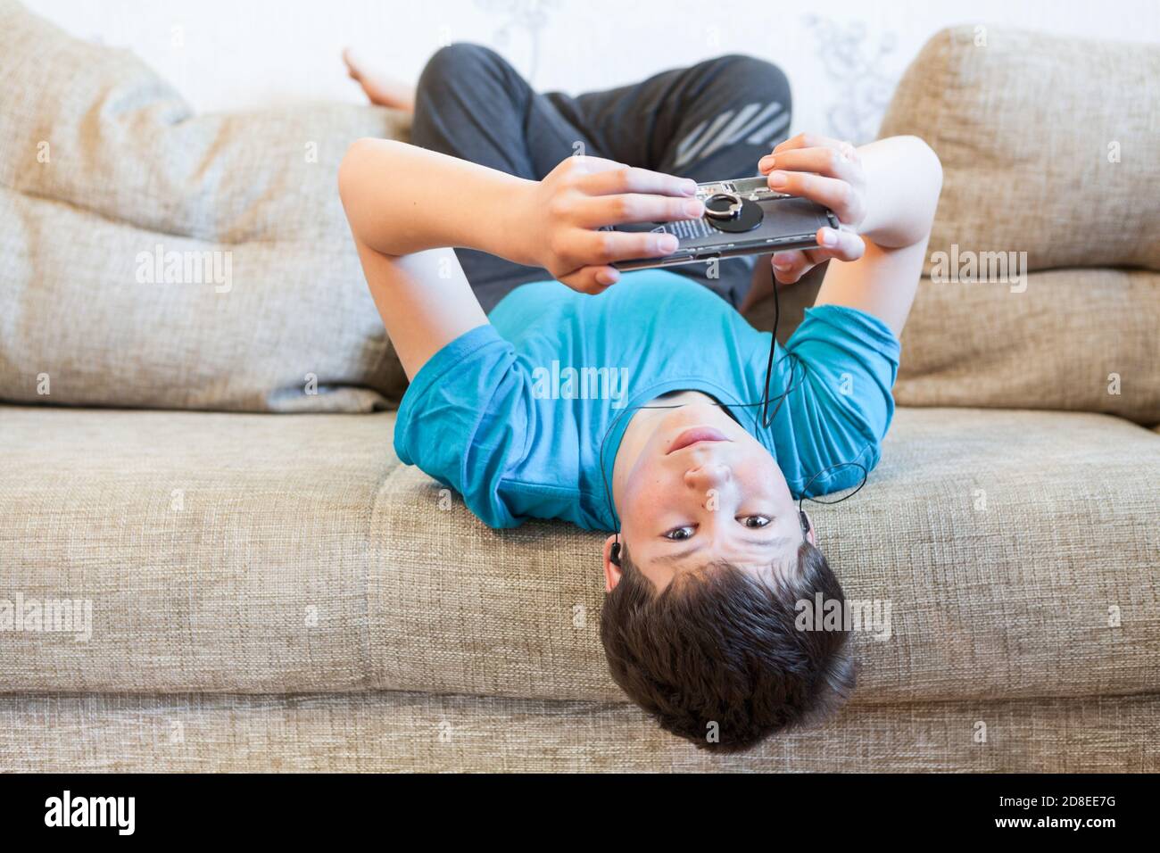 Attraktiver Jugendlicher in blauem T-Shirt auf dem Rücken auf der Couch liegend und Smartphone in den Händen haltend, in sozialen Medien surfen Stockfoto