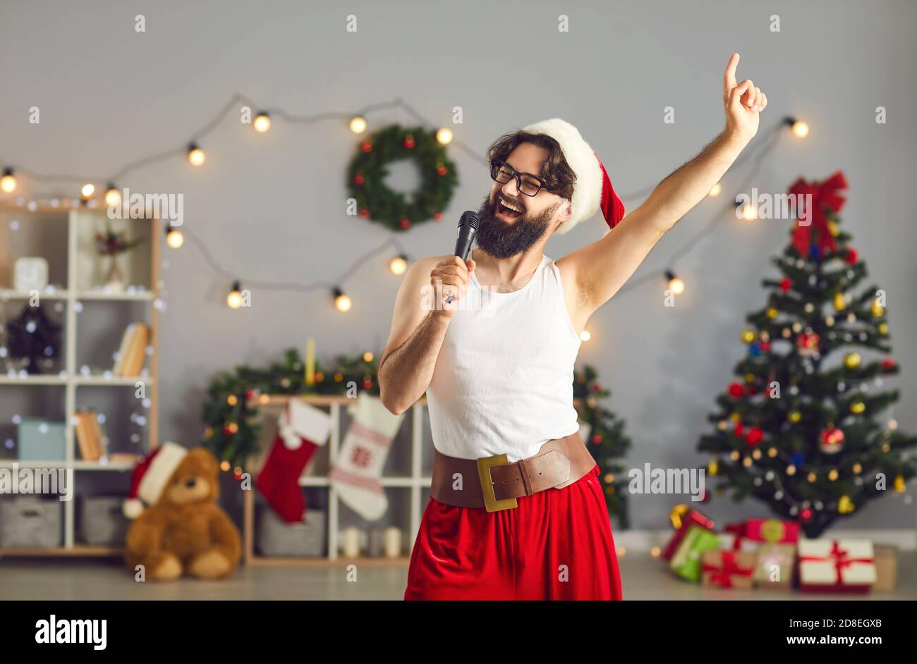 Lustig lächelnder Mann Santa in festlicher Heimkleidung feiert Weihnachten Und zu Hause im Mikrofon singen Stockfoto