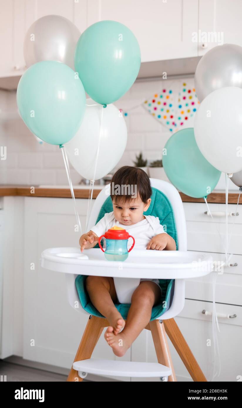 Kleiner Junge sitzt im Hochstuhl zu Hause auf Weiße Küche und Trinkwasser aus sippy Tasse auf Hintergrund Mit Luftballons Stockfoto