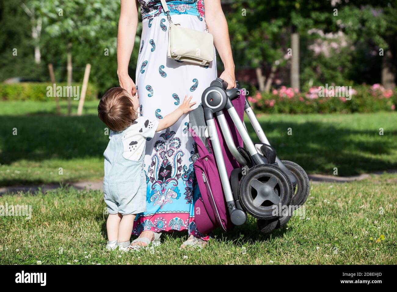 Launisch Kind bittet seine Mutter, in den Armen zu nehmen, unkenntlich Frau hält Baby Kinderwagen in der Hand, Sommerwiese im sonnigen Park Stockfoto