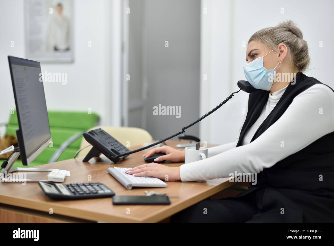 Junge Verkaufsleiterin in einer Gesichtsmaske im Gespräch Das Telefon im Büro Stockfoto
